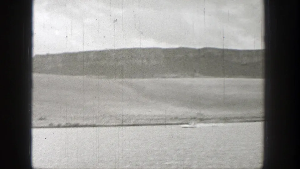 1938 Washington home-movie frame with a small boat near the bottom edge, calm water, and a long low hillside beyond.