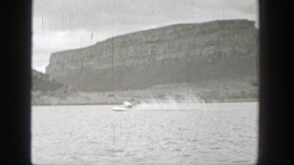 1938 Washington 8mm frame with a small boat on rippled lake water and a broad rocky hillside in the background.