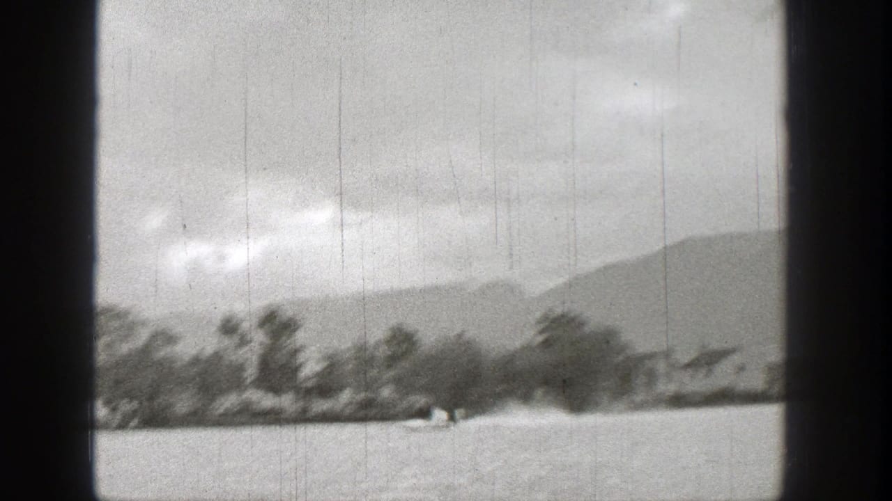Small motorized boat crossing a lake near trees and distant hills under a cloudy sky.