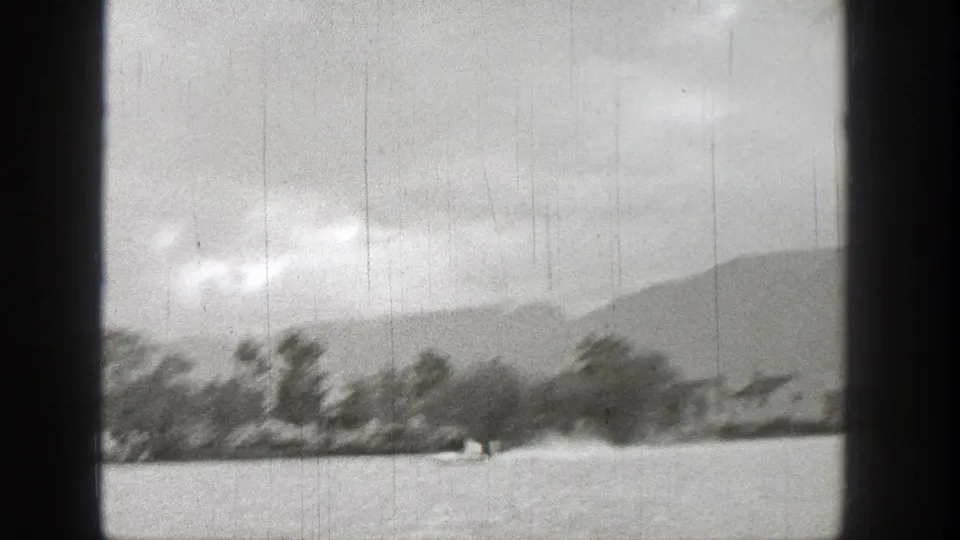 1938 Washington 8mm frame showing a small fast watercraft moving across a lake with tree-lined shore and distant hills.