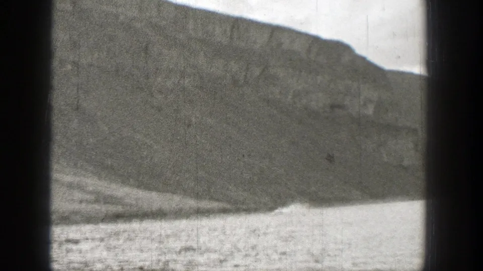 The frame shows a steep rocky slope meeting a lake, with soft focus, grain, and no identifiable people or structures.