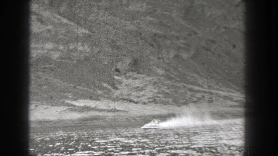 Archival 8mm frame of a motorboat throwing spray as it passes below a steep Washington lakeside slope.
