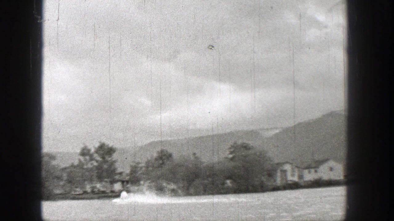 Small motorboat and wake near a tree-lined shore with houses and hills in the background.