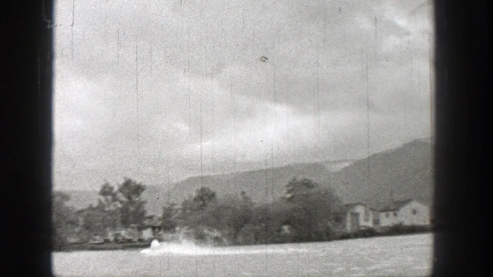 Black-and-white 8mm view of a small motorboat moving past a Washington lakeshore with houses, trees, and hills.