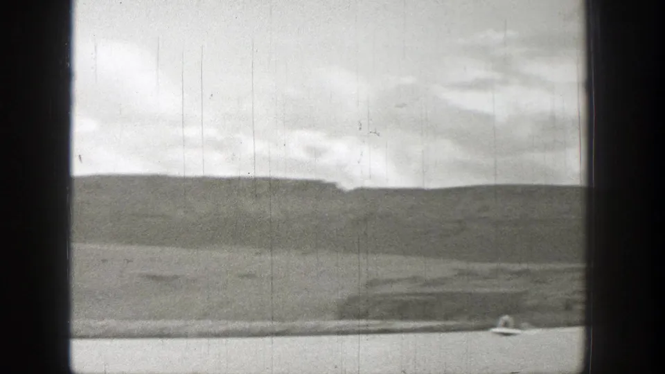 Wide black-and-white 8mm frame of a distant motorboat on a Washington lake below rolling hills and a cloudy sky.