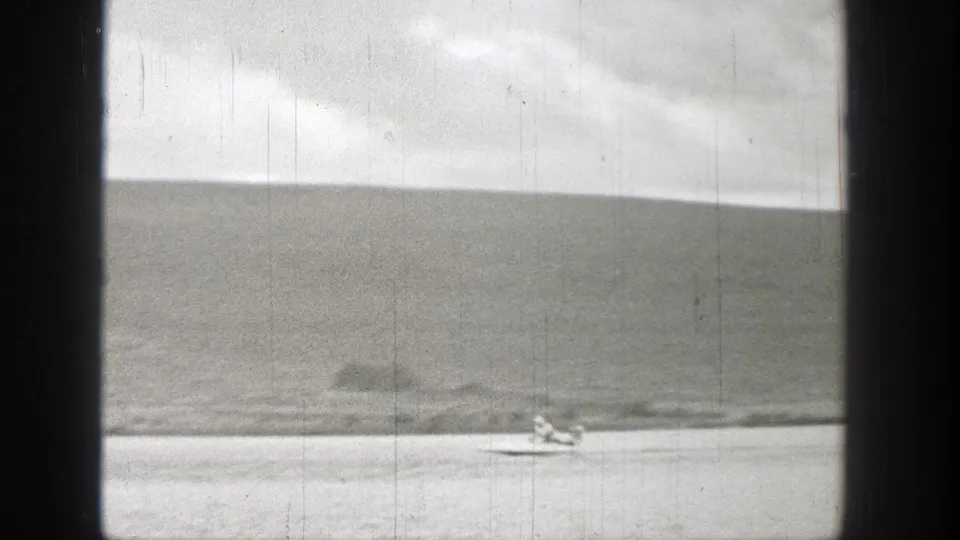 A small watercraft moves past a stationary object near the Washington riverbank in a 1938 8mm frame.