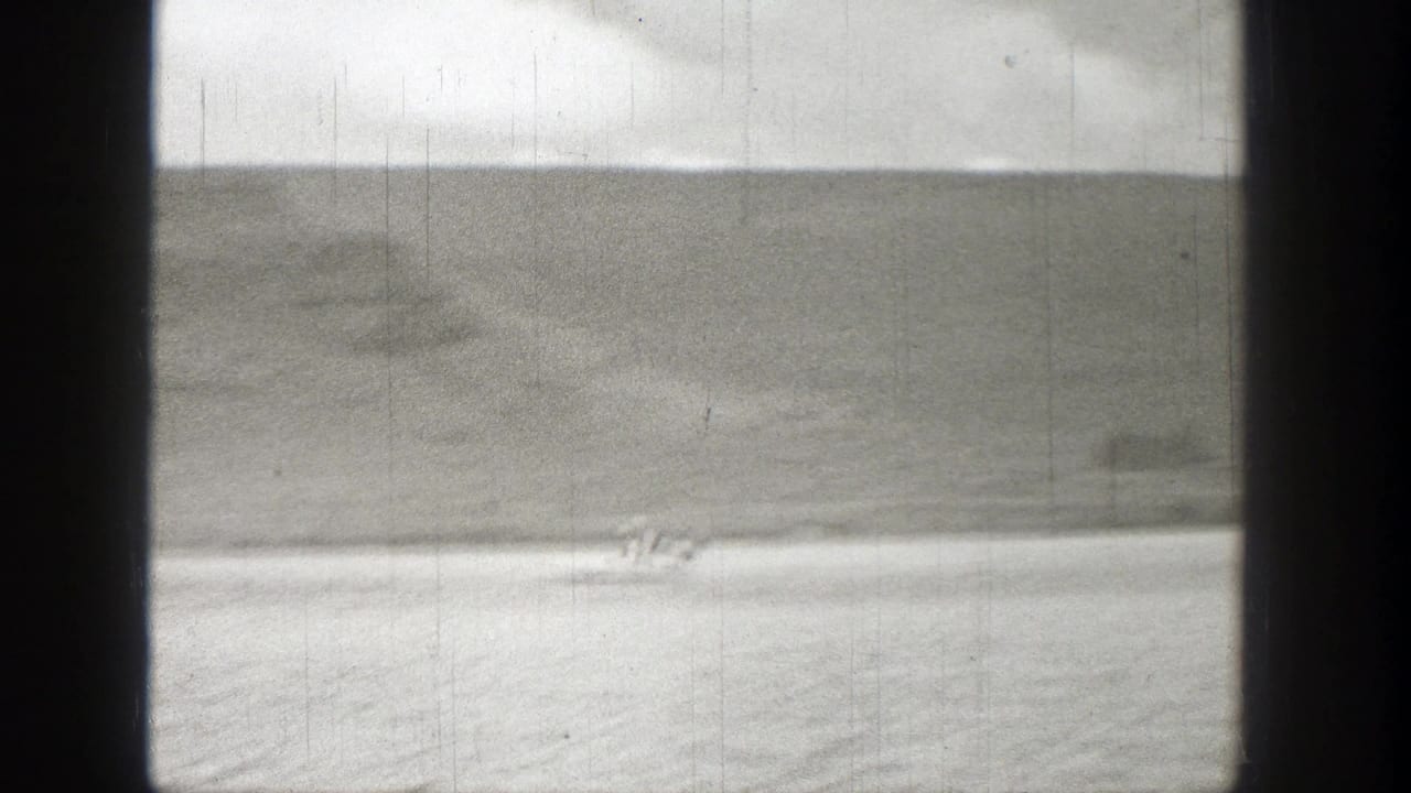 1938 Washington 8mm frame showing a small distant craft or rider on water near a pale shore and rugged hillside.