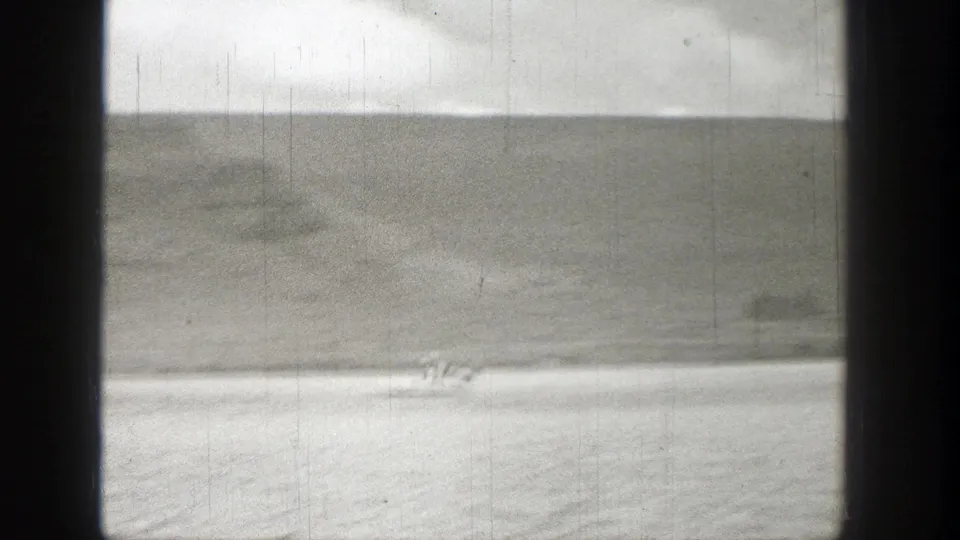 1938 Washington 8mm frame showing a small distant craft or rider on water near a pale shore and rugged hillside.