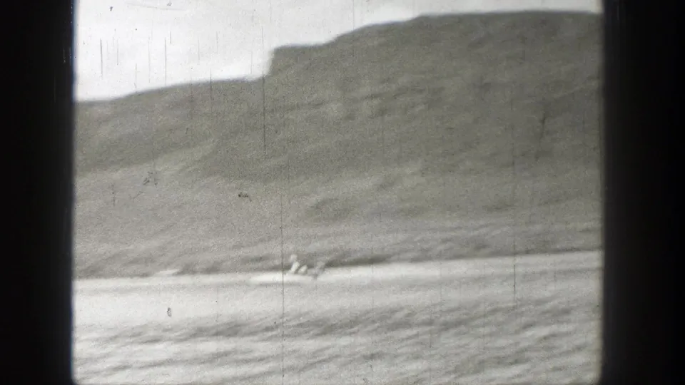 A distant figure or low craft appears on the water near a Washington shoreline, framed by soft 8mm grain and dark borders.