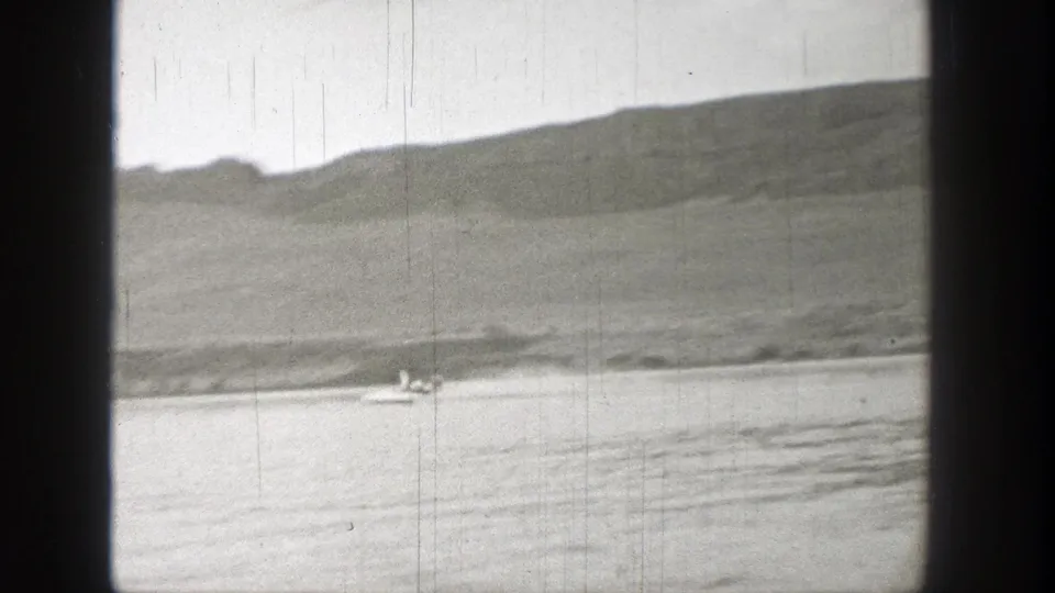 Black-and-white 8mm view of a small low craft near shore, with ripples and a rugged Washington hillside in the background.
