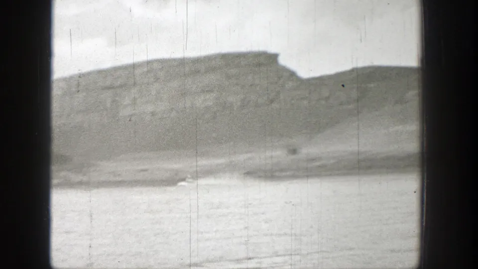 1938 Washington 8mm frame showing a broad lake surface below a soft, rocky mountain ridge under cloudy sky.