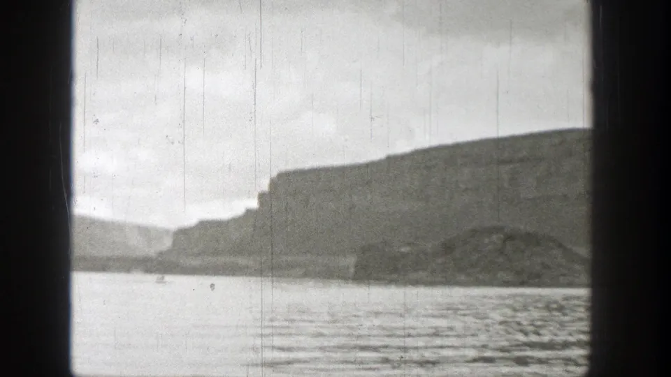 1938 Washington 8mm frame showing rippled water and a distant flat-topped cliff or bluff beneath an overcast sky.