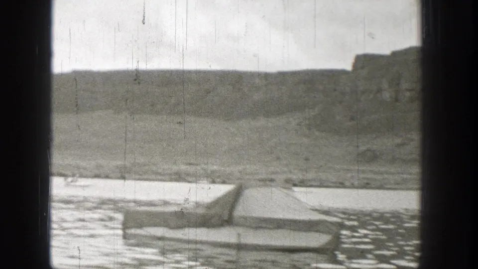 1938 Washington 8mm frame with dark window bars, water, shoreline, and a low vehicle-like form against distant hills.