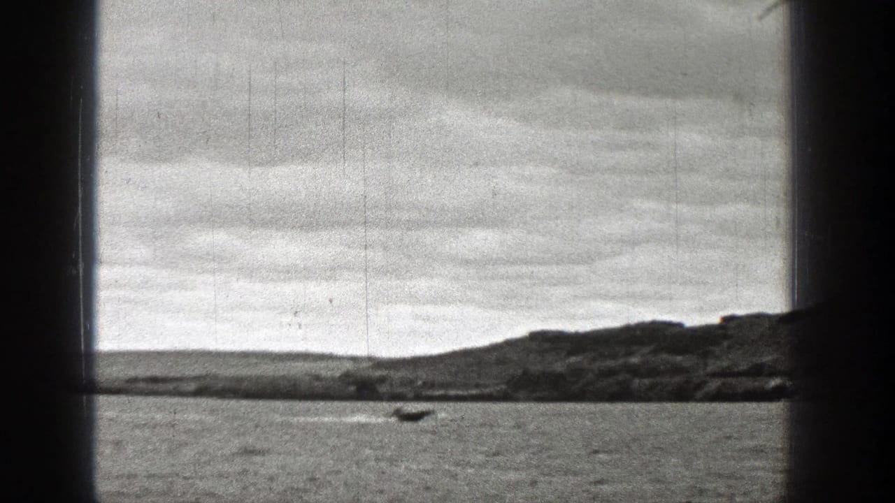 Black-and-white 8mm frame showing open water, a small boat, and a low Washington shoreline beneath an overcast sky.