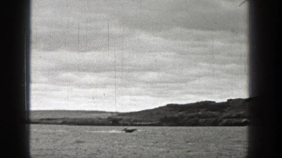 Black-and-white 8mm frame showing open water, a small boat, and a low Washington shoreline beneath an overcast sky.