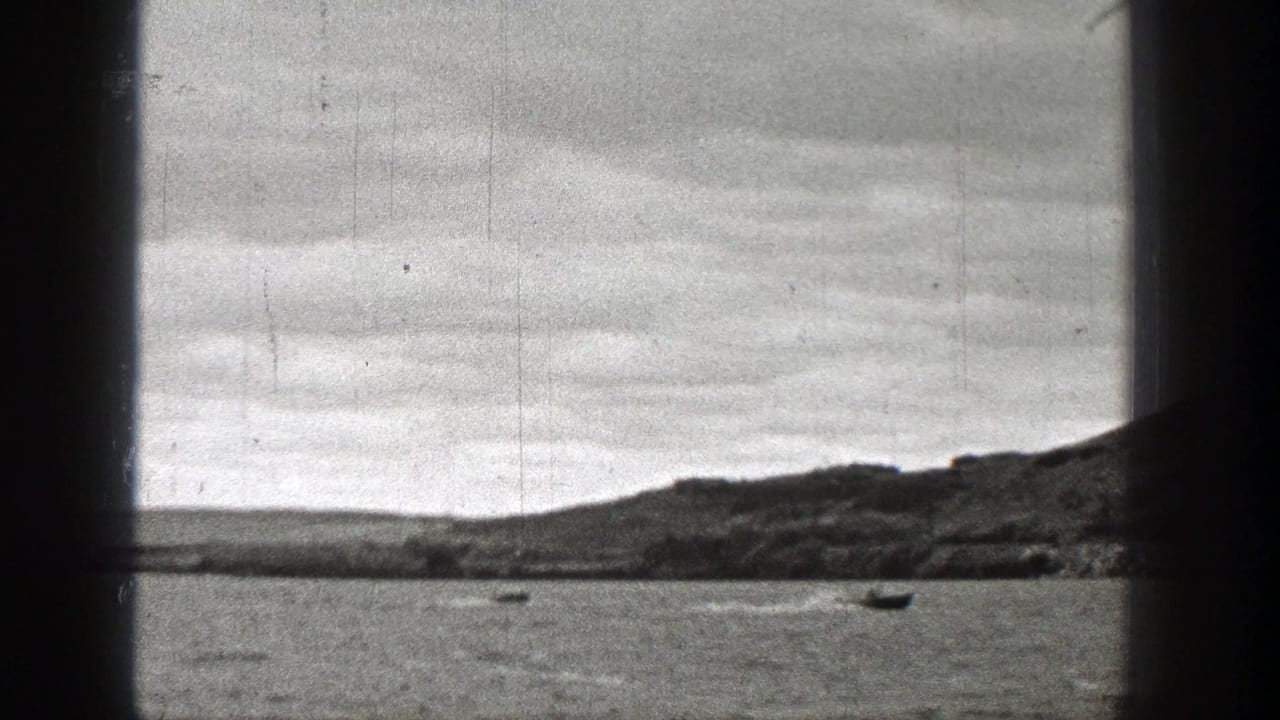 1938 Washington home-movie frame with a vertical window mullion, small boats on water, a low shoreline, and overcast sky.