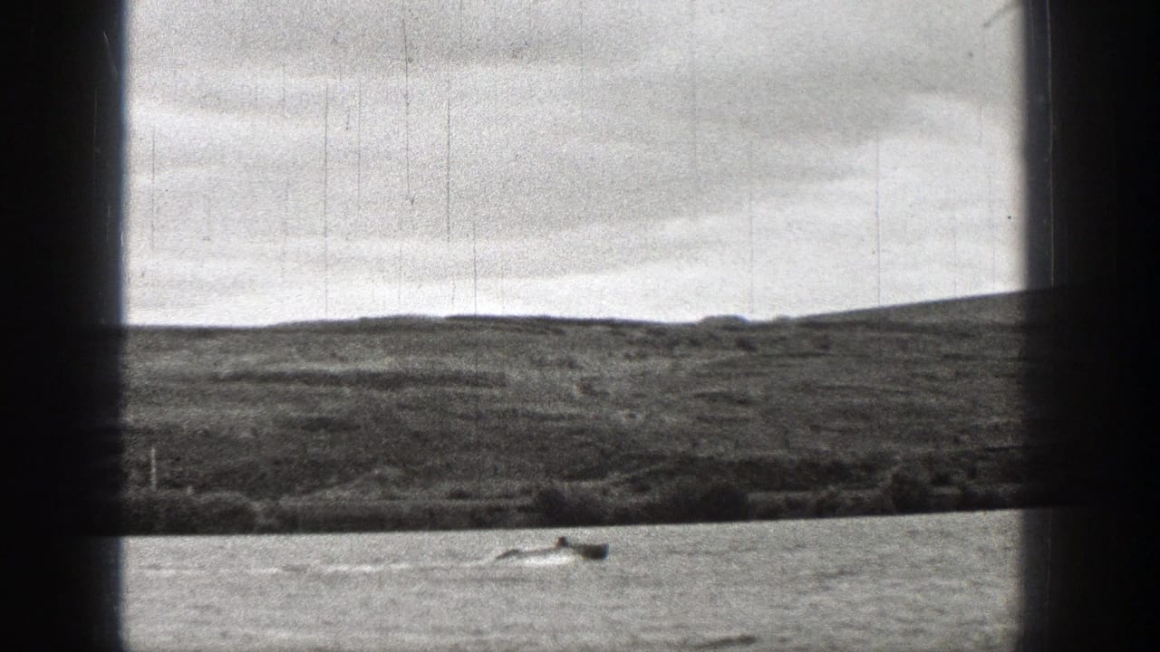 1938 Washington 8mm view through vessel window bars, showing rippled water, a small boat, low grassy land, and a cloudy sky.