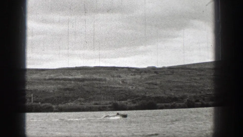 1938 Washington 8mm view through vessel window bars, showing rippled water, a small boat, low grassy land, and a cloudy sky.