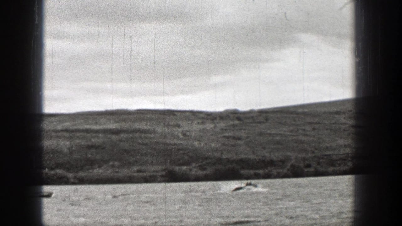 1938 Washington frame showing a small boat near the lower edge, rippled water, low shoreline, dark window framing, and overcast sky.
