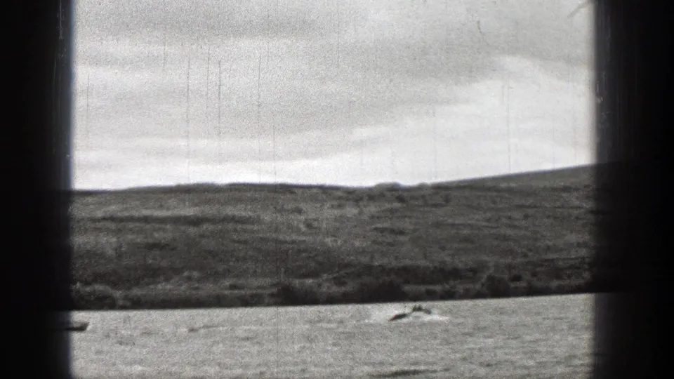 1938 Washington frame showing a small boat near the lower edge, rippled water, low shoreline, dark window framing, and overcast sky.
