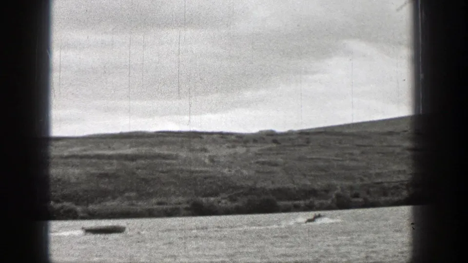 1938 Washington 8mm frame showing a small boat moving across a lake below grassy hills and an overcast sky.