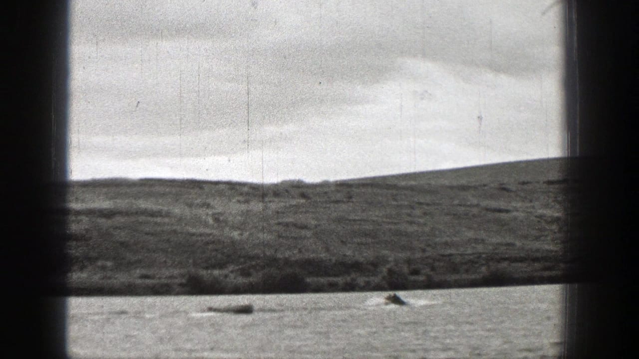 1938 Washington lakeside frame with a distant boat and wake against a grassy field and rolling hill.