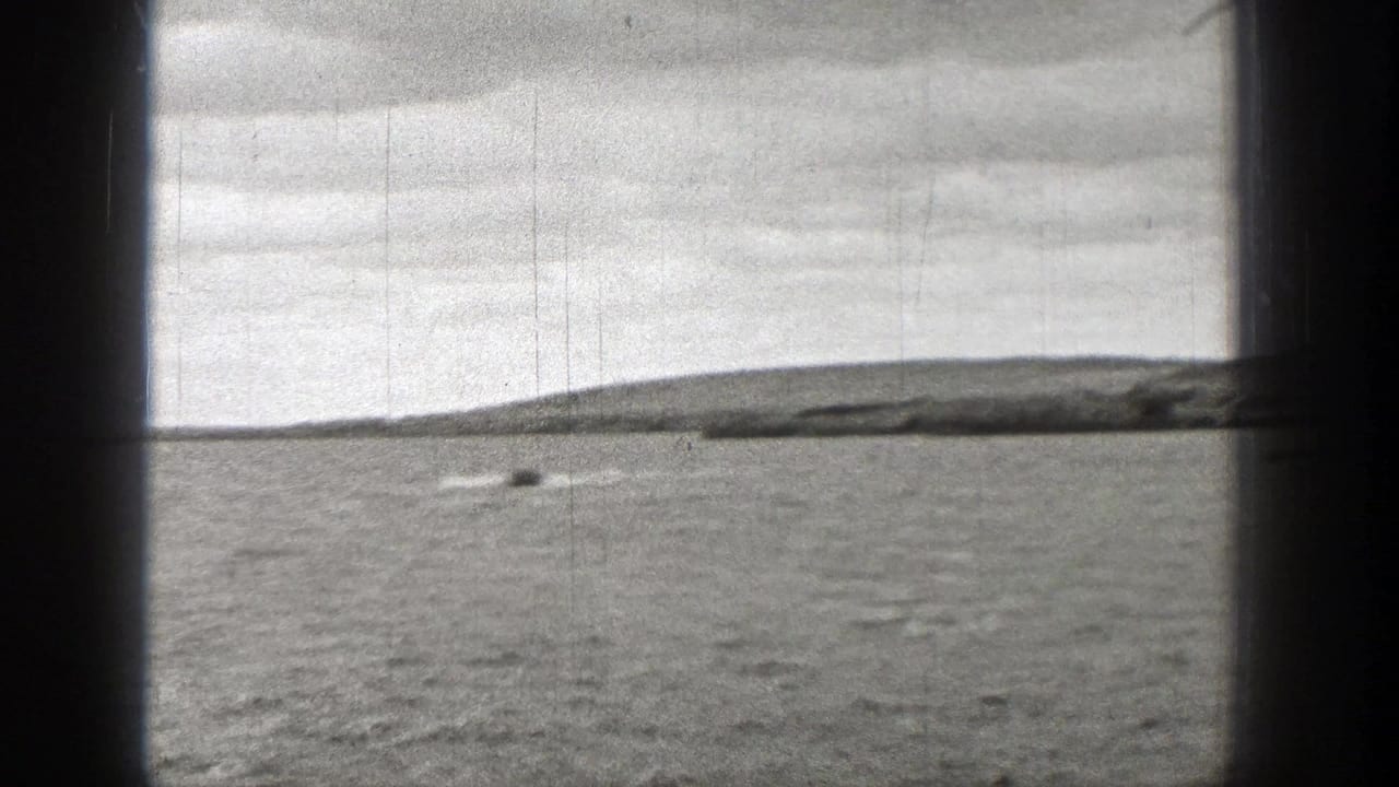1938 Washington 8mm frame showing a small boat and wake across rippled water, with a low shoreline and overcast sky in the distance.