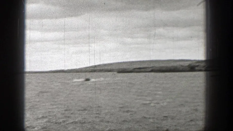 1938 Washington 8mm frame showing a small boat and wake across rippled water, with a low shoreline and overcast sky in the distance.
