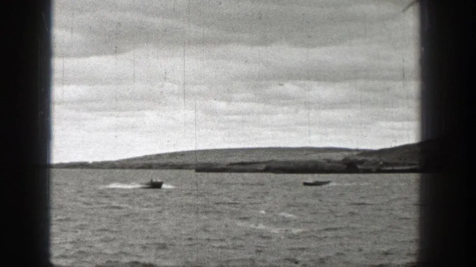 1938 Washington frame with two small boats moving across choppy water, their wakes visible below a low hilly shoreline and cloudy sky.