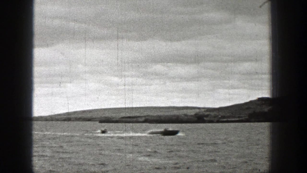 1938 Washington 8mm frame with a speedboat cutting across lake water, another boat nearby, low hills, cloudy sky, and scratched film texture.