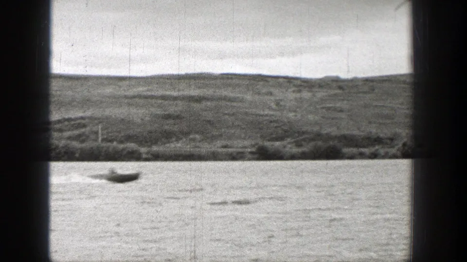 1938 Washington home-movie frame showing a small boat on open water below a low, rugged shoreline.