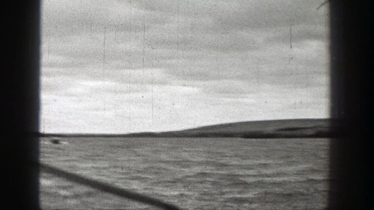 Grainy 1938 Washington coastal frame showing ocean water, a low island-like silhouette, and cloudy sky.