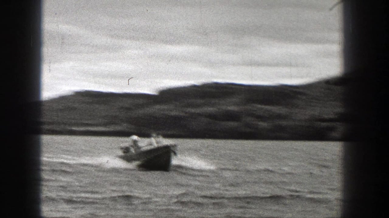 1938 Washington 8mm frame with a small open motorboat nearer the camera, tiny occupants visible, and a low wooded shoreline behind.