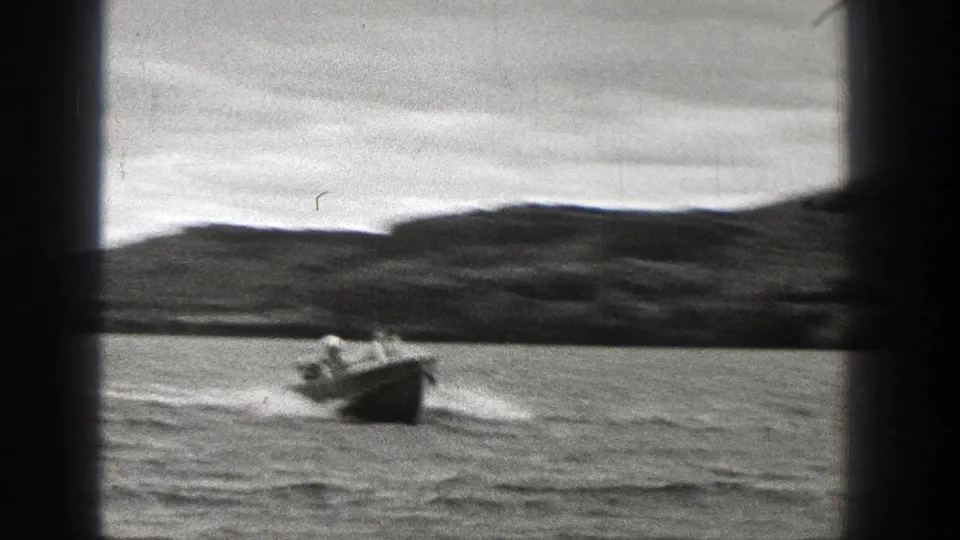 1938 Washington 8mm frame with a small open motorboat nearer the camera, tiny occupants visible, and a low wooded shoreline behind.