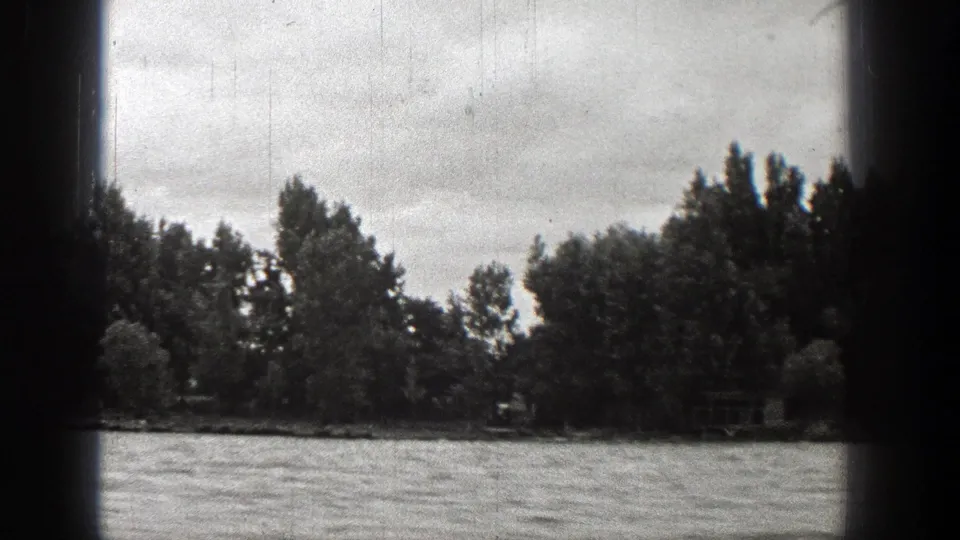 1938 Washington lake scene with rippled water, wooded shoreline, cloudy sky, and visible 8mm film scratches.