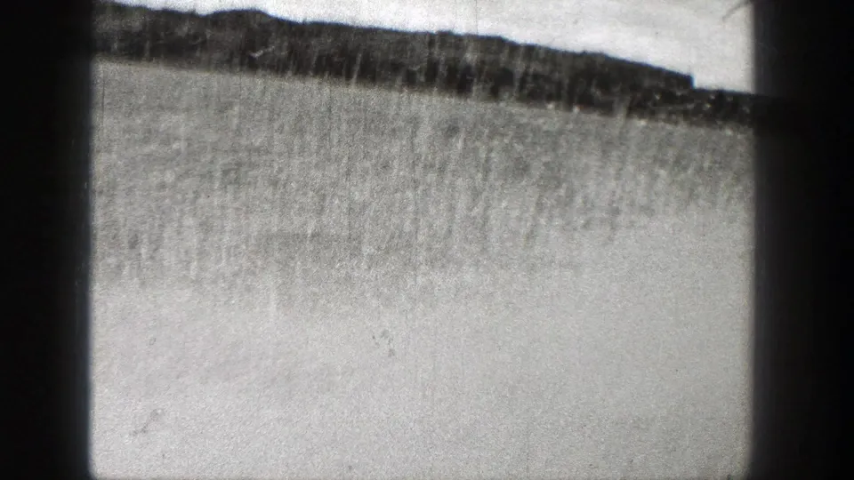 Black-and-white 8mm frame of a low Washington shoreline across bright water, with scratches and dark border shadows.