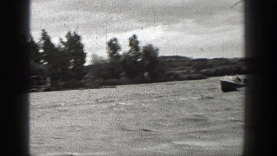 1938 Washington 8mm frame of a small motorboat near a tree-lined lakeshore, with people faintly visible aboard.