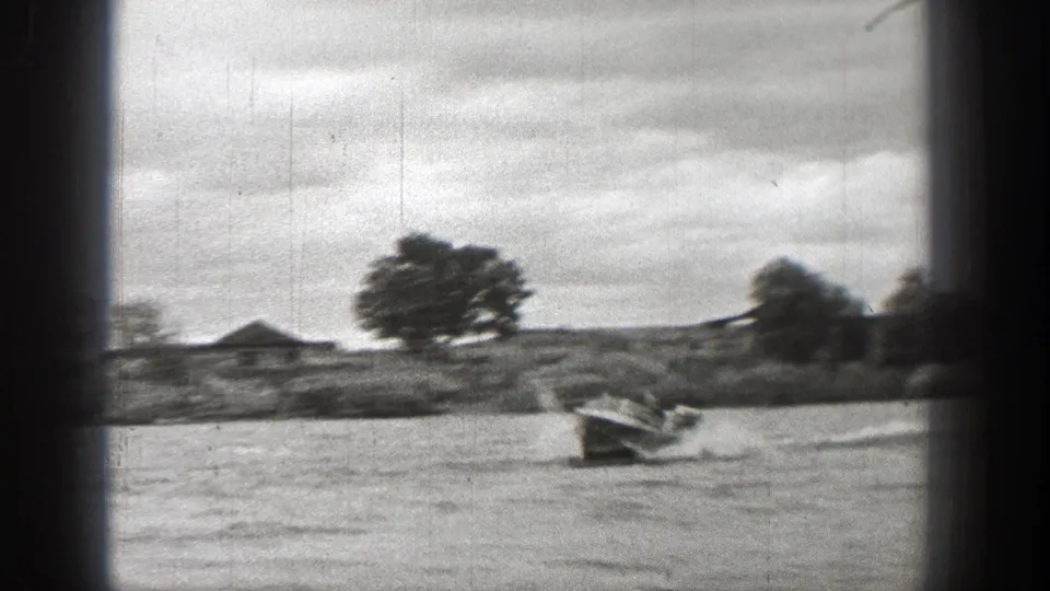 1938 Washington 8mm frame of a small motorboat on lake water near an open shoreline with trees and a distant structure.