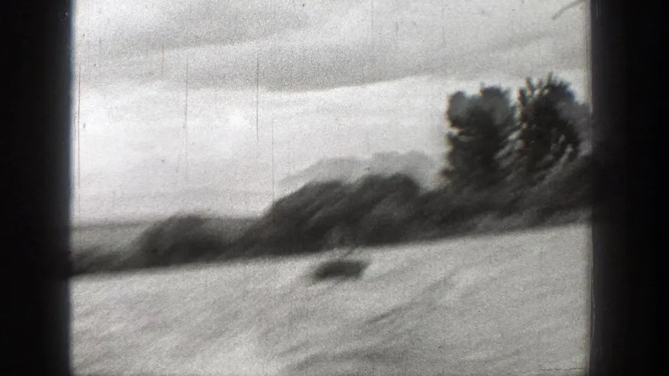 1938 Washington 8mm frame with blurred water, low shoreline vegetation, trees, cloudy sky, and vertical film scratches.