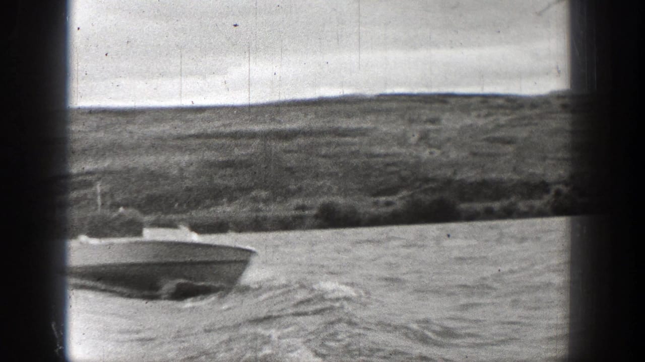 Washington 1938 home-movie frame showing a small speedboat crossing choppy water near a low shoreline, with dark film borders visible.