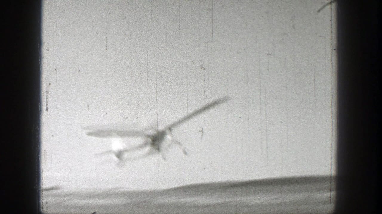 Small blurred airplane crossing a pale sky with dark film-frame edges.
