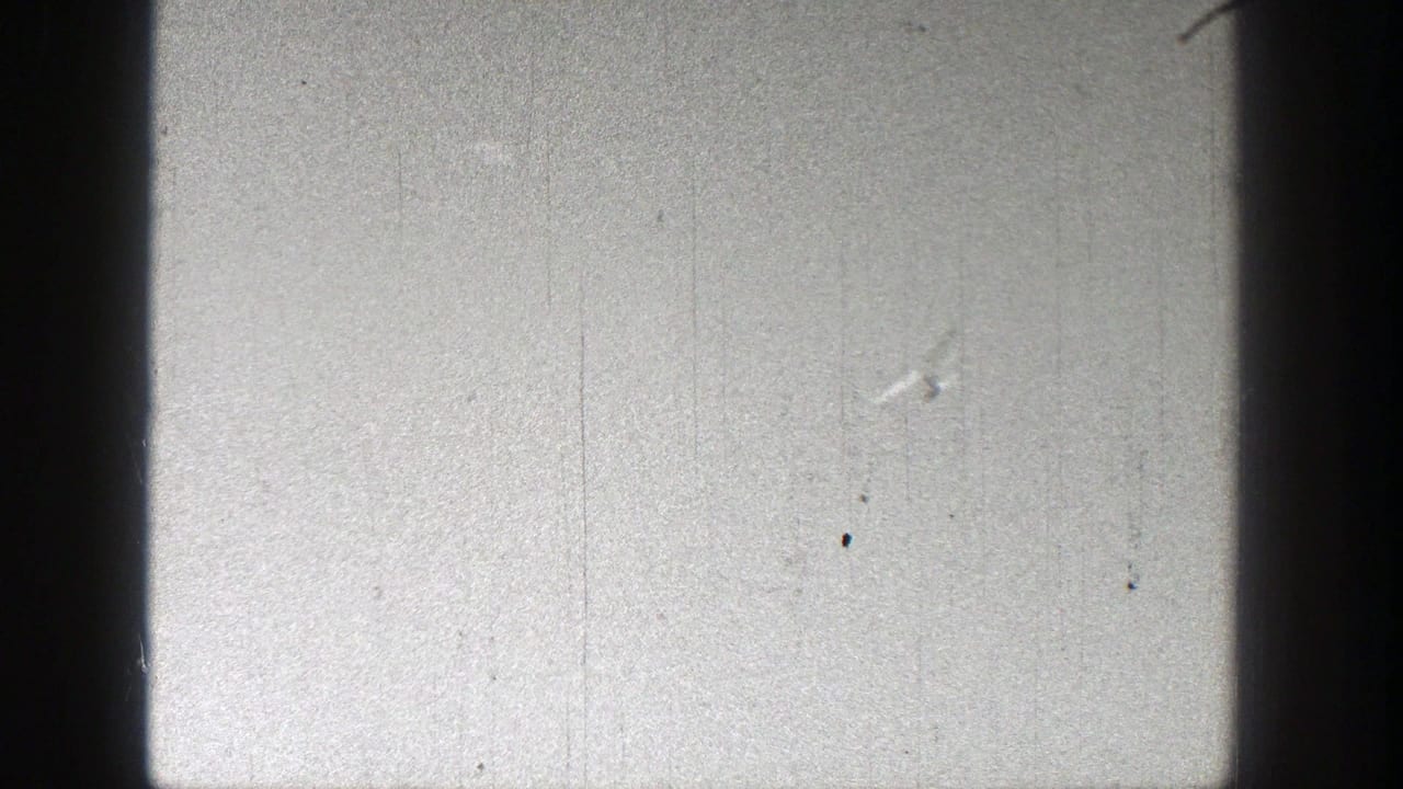 Tiny pale model airplane against a bright gray sky with film grain and scratches.
