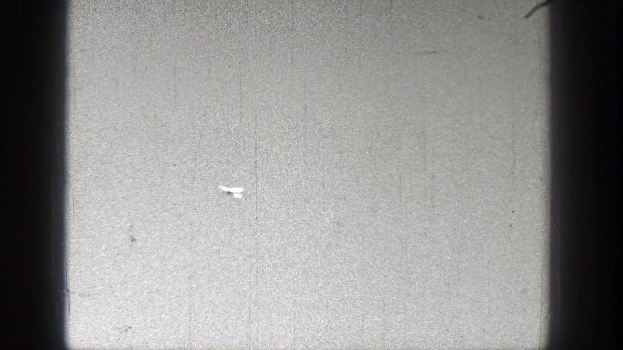 Small white model airplane crossing a pale sky with scratches and dust on the film.