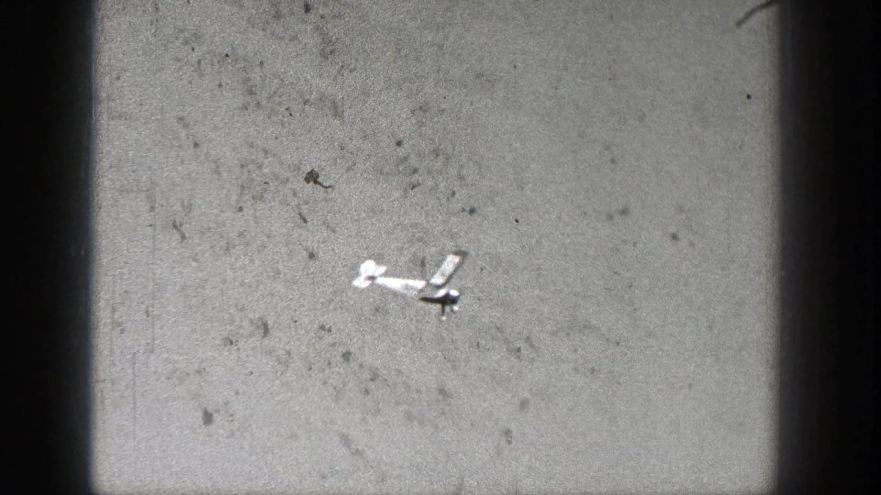 A small model aircraft appears in flight against a pale sky in 1938 Washington home-movie footage, with visible grain and frame edge shadowing.