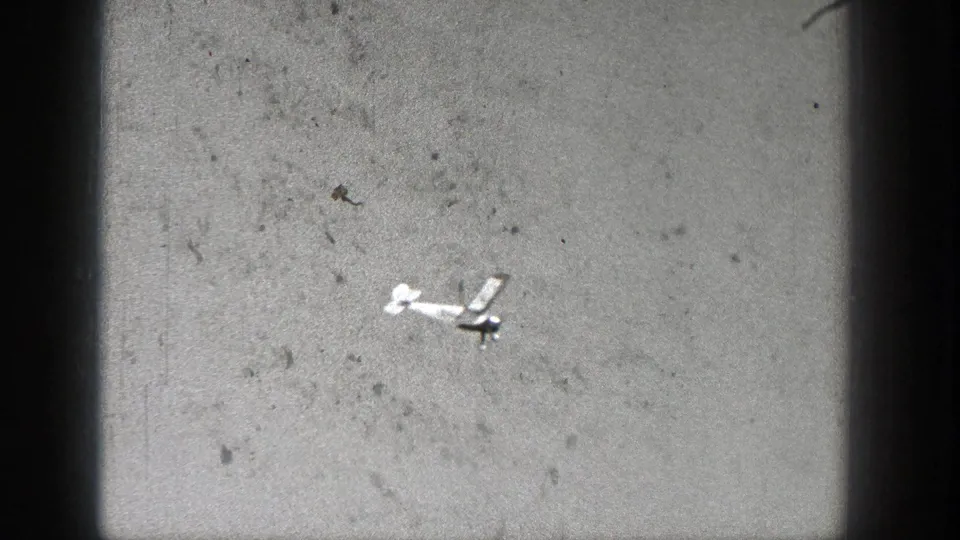 A small model aircraft appears in flight against a pale sky in 1938 Washington home-movie footage, with visible grain and frame edge shadowing.