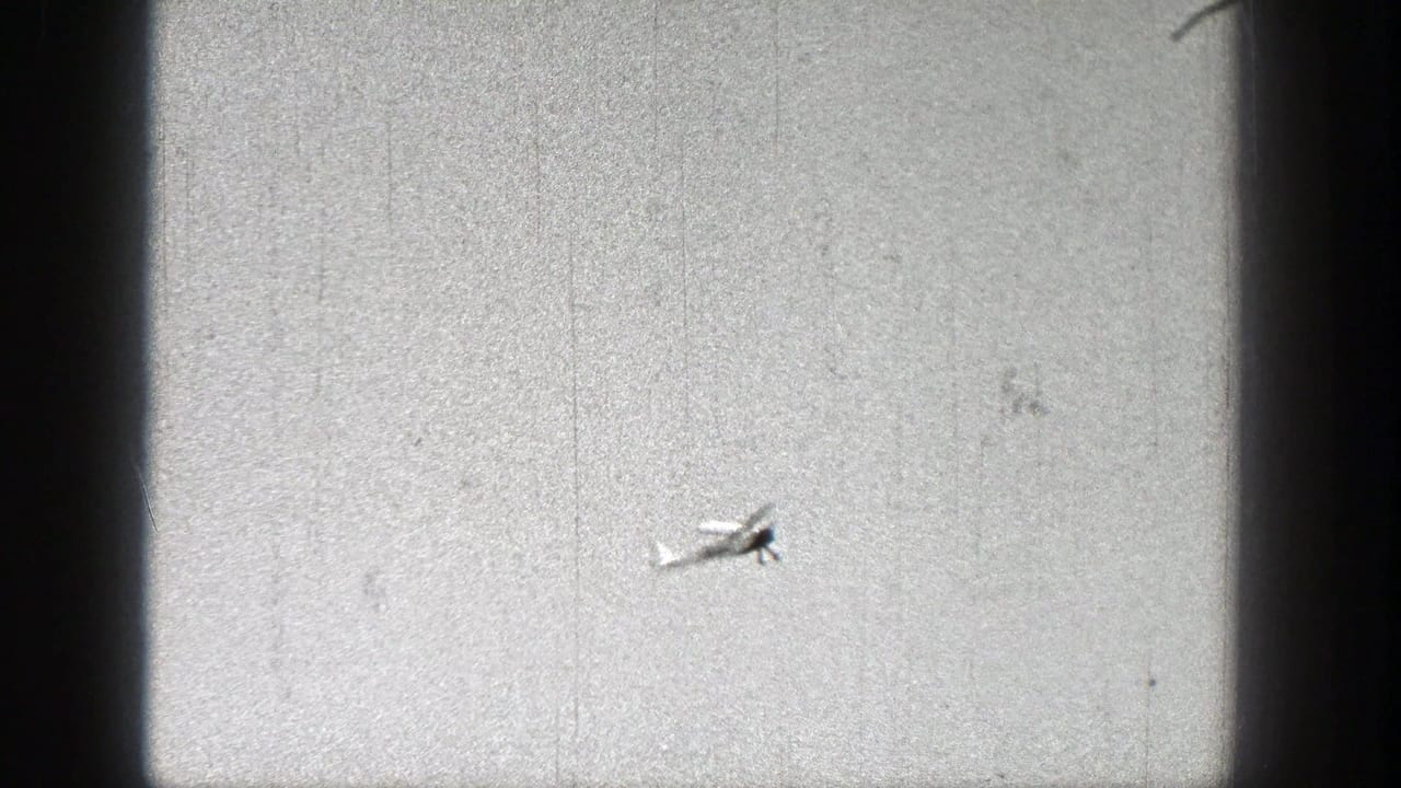 Small model airplane flying across a bright, grainy sky.