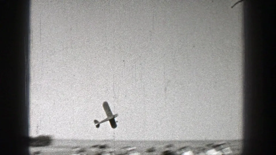 The model aircraft banks above a flat open landscape in the supplied 1938 Washington home movie, framed by dark 8mm edge shadows.