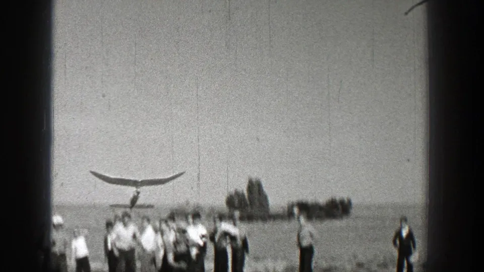 A glider-like object appears above distant people near a waterline or open field in 1938 Washington home-movie footage.