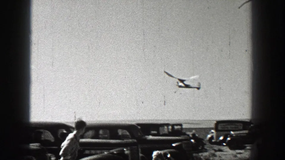 Washington 1938 frame with spectators near parked cars while a small plane flies above the horizon.