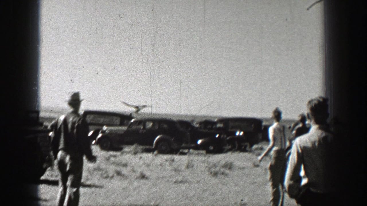 Men stand in a dusty field near parked cars as a small model airplane flies overhead.
